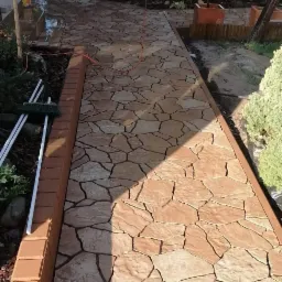 Ścieżka z nieregularnych, brązowo-beżowych kamieni ułożona w ogrodzie, obrzeża z cegieł, widoczny grill murowany w tle.