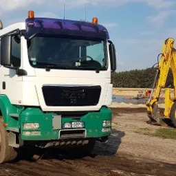 Ciężarówka MAN z zieloną skrzynią ładunkową i pies obok, w tle żółta koparko-ładowarka New Holland, na terenie wyrobiska.