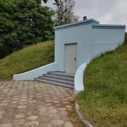 Niebieski budynek z płaskim dachem, częściowo wkomponowany w zielony pagórek, z szarymi schodami i chodnikiem z betonowej kostki.