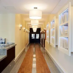 Długi korytarz z drewnianą podłogą i oświetleniem w postaci wiszących lamp cylindrycznych oraz kinkietów, dekorowany wnękami z książkami podświetlonymi LED i fragmentem ściany z czerwonej cegły.