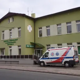 Budynek szpitala w odcieniach zieleni z białymi akcentami, z zaparkowaną karetką pogotowia na pierwszym planie, widoczny napis 'Szpital Zespolony w Kędzierzynie-Koźlu Budynek B' i symbol medyczny.