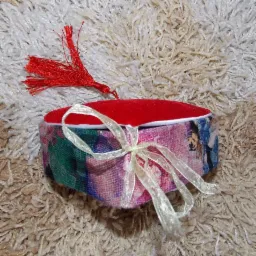 Ozdobne pudełko na biżuterię z czerwonym, aksamitnym wieczkiem, wzorzystym bokiem i kremową wstążką, leżące na puszystym dywanie.