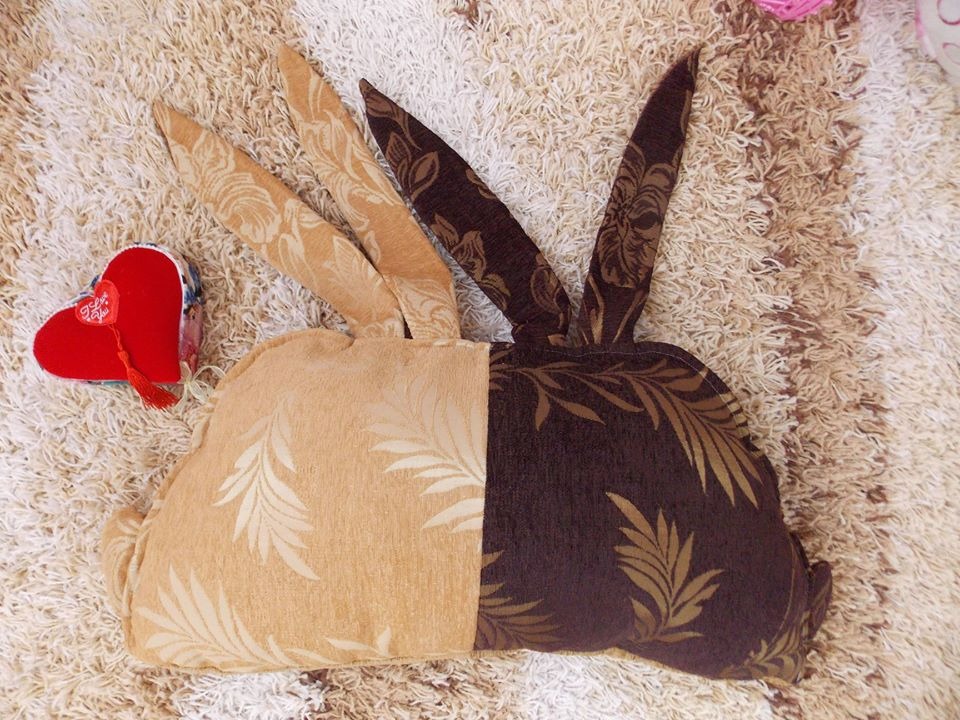 Dwukolorowa poduszka w kształcie królika z uszami, wykonana z tkaniny w liście, leżąca na puszystym dywanie obok czerwonego serduszka.