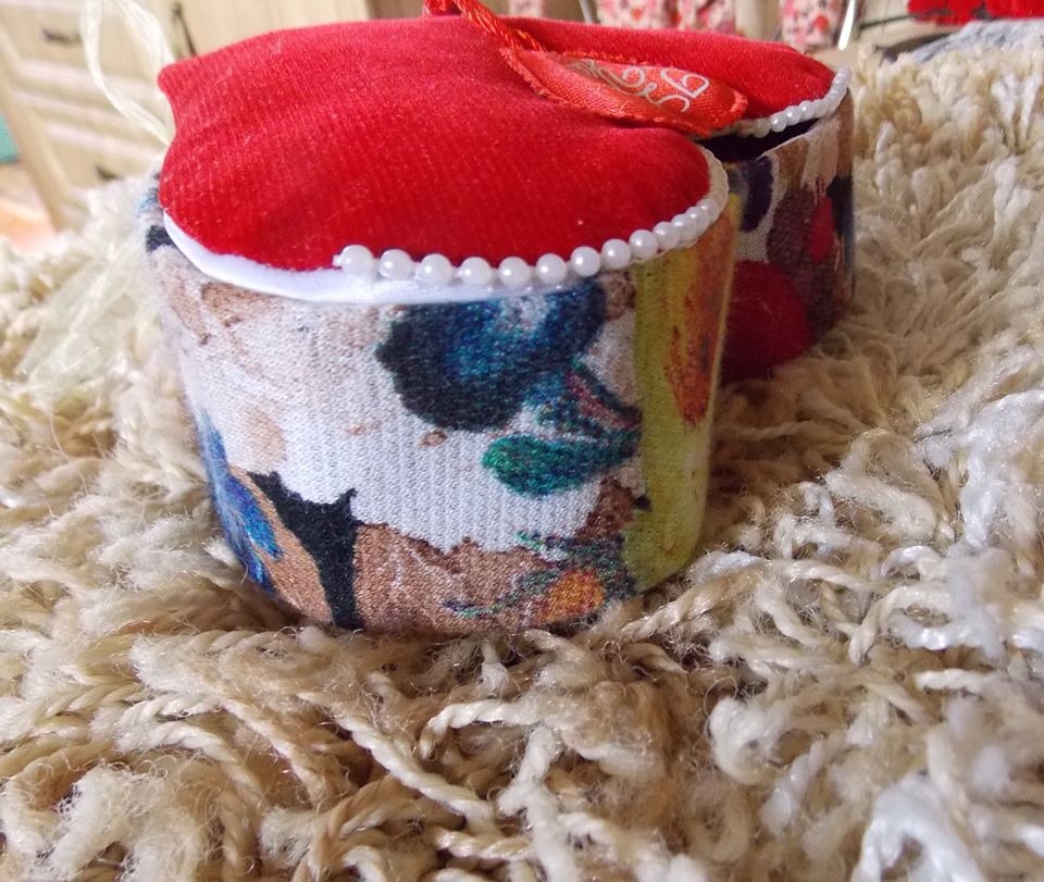 Dwa małe, ręcznie robione pufy z czerwonymi, welurowymi siedziskami ozdobionymi białymi koralikami, na wzorzystych, materiałowych podstawach, leżące na puszystym, kremowym dywanie.