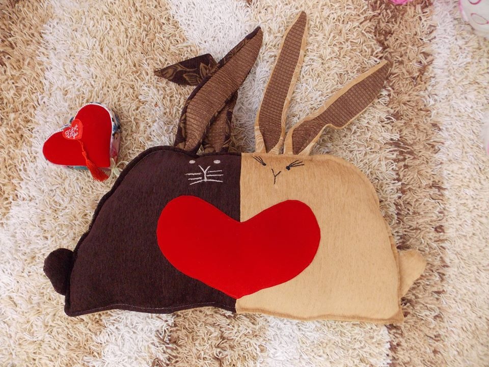 Dwukolorowa poduszka w kształcie zająca, z brązową i beżową połową, ozdobiona czerwonym sercem, leżąca na puszystym dywanie obok małego czerwonego pudełka w kształcie serca.