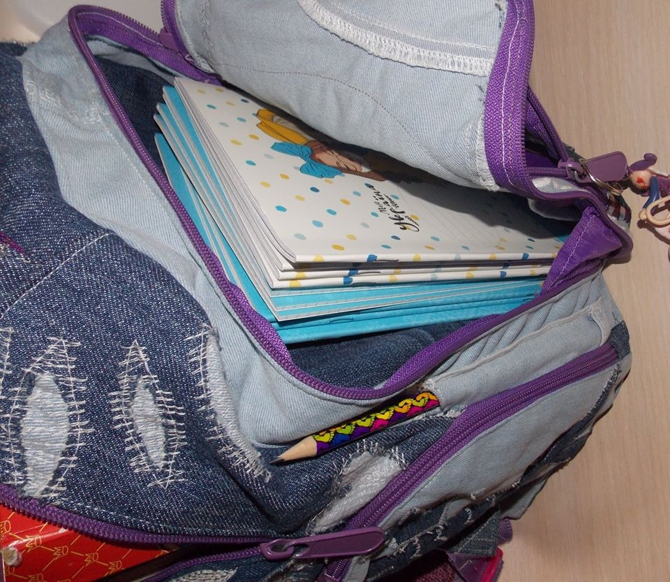 Plecak wykonany z przetworzonego denimu w odcieniach niebieskiego, z fioletowymi zamkami błyskawicznymi i wzorem haftu, wypełniony zeszytami i kredką w bocznej kieszeni.