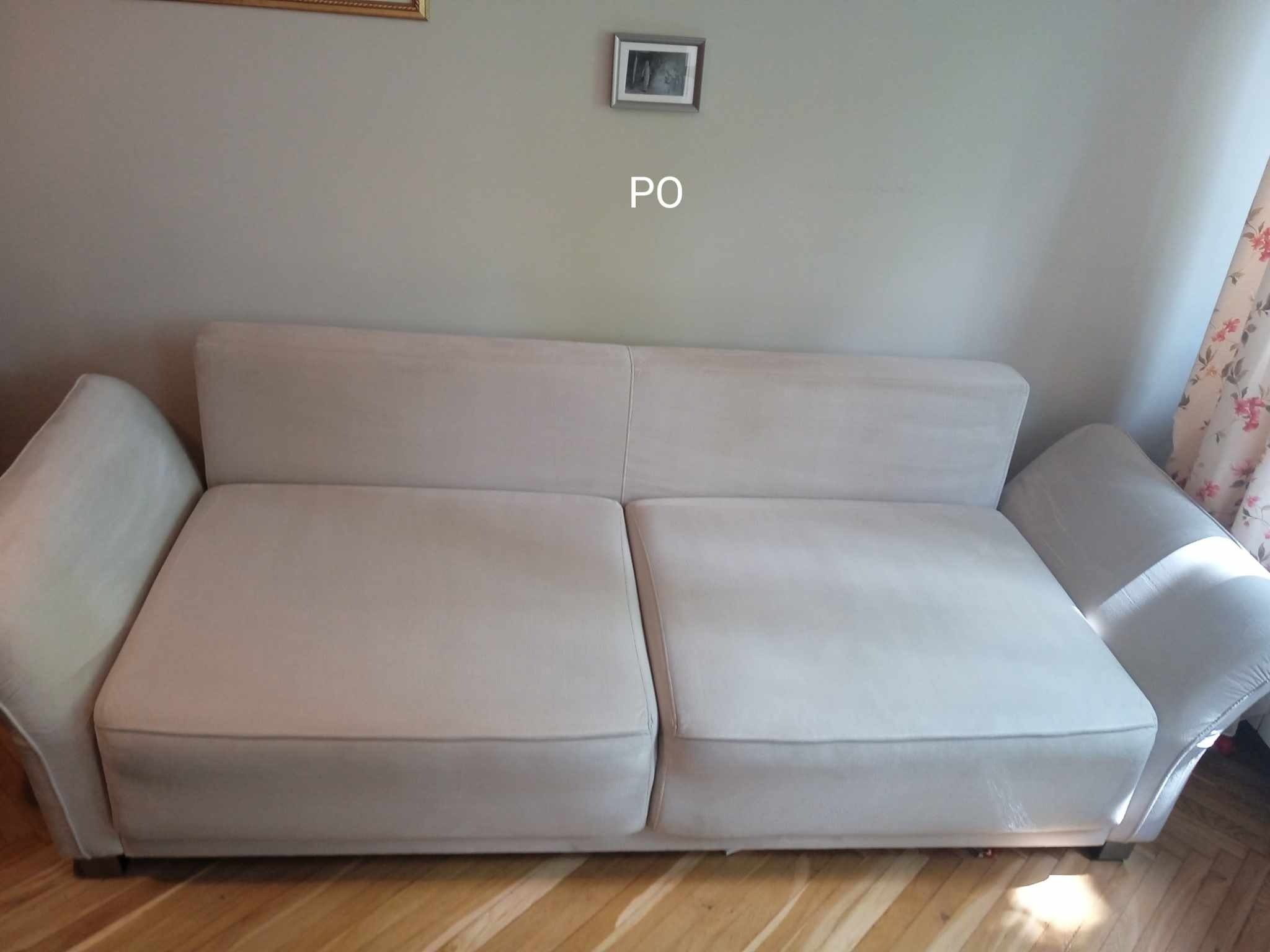 Jasnoszara kanapa dwuosobowa po czyszczeniu, widoczne delikatne ślady wilgoci na tapicerce, drewniana podłoga, ściana z obrazkiem i zasłoną.