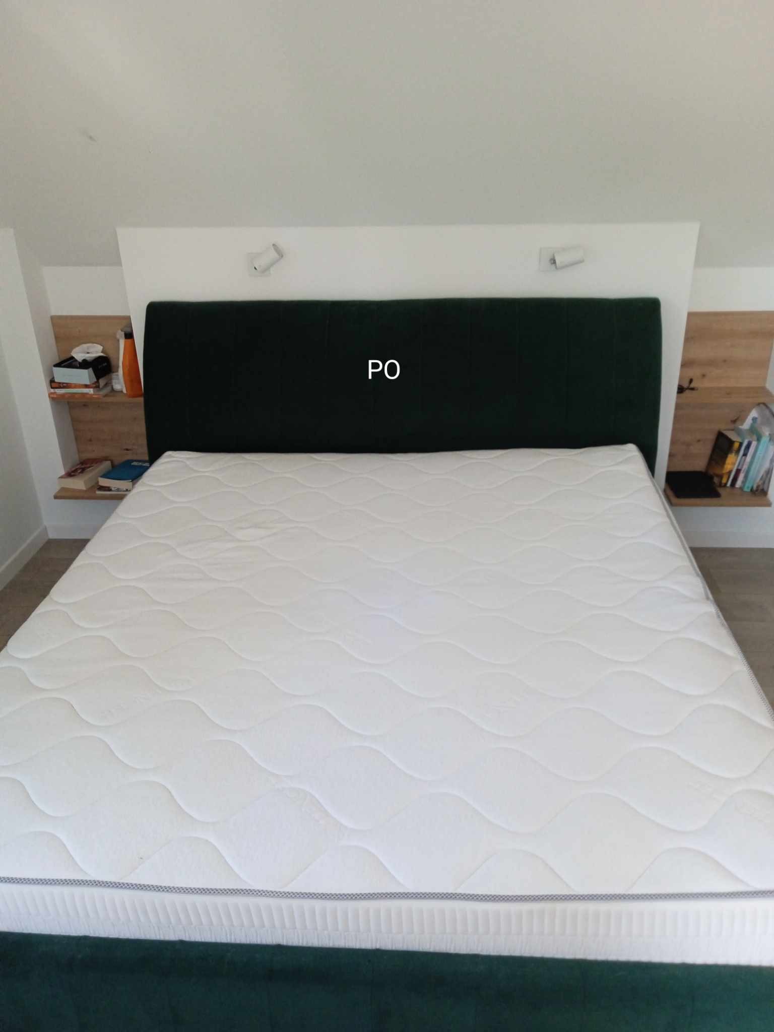 Nowy materac na łóżku z ciemnozielonym zagłówkiem, z półkami nocnymi po bokach, oświetlonymi lampkami.