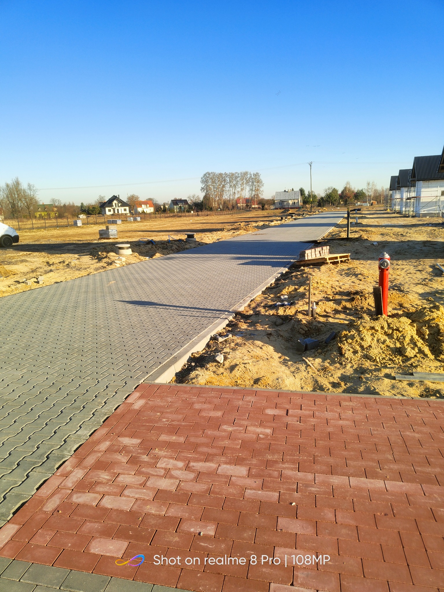 Fragment nowo wyłożonego chodnika z kostki brukowej w dwóch kolorach: szarym i ceglastym, na tle piaszczystego terenu budowy z hydrantem i częściowo wykończonymi domami w oddali.