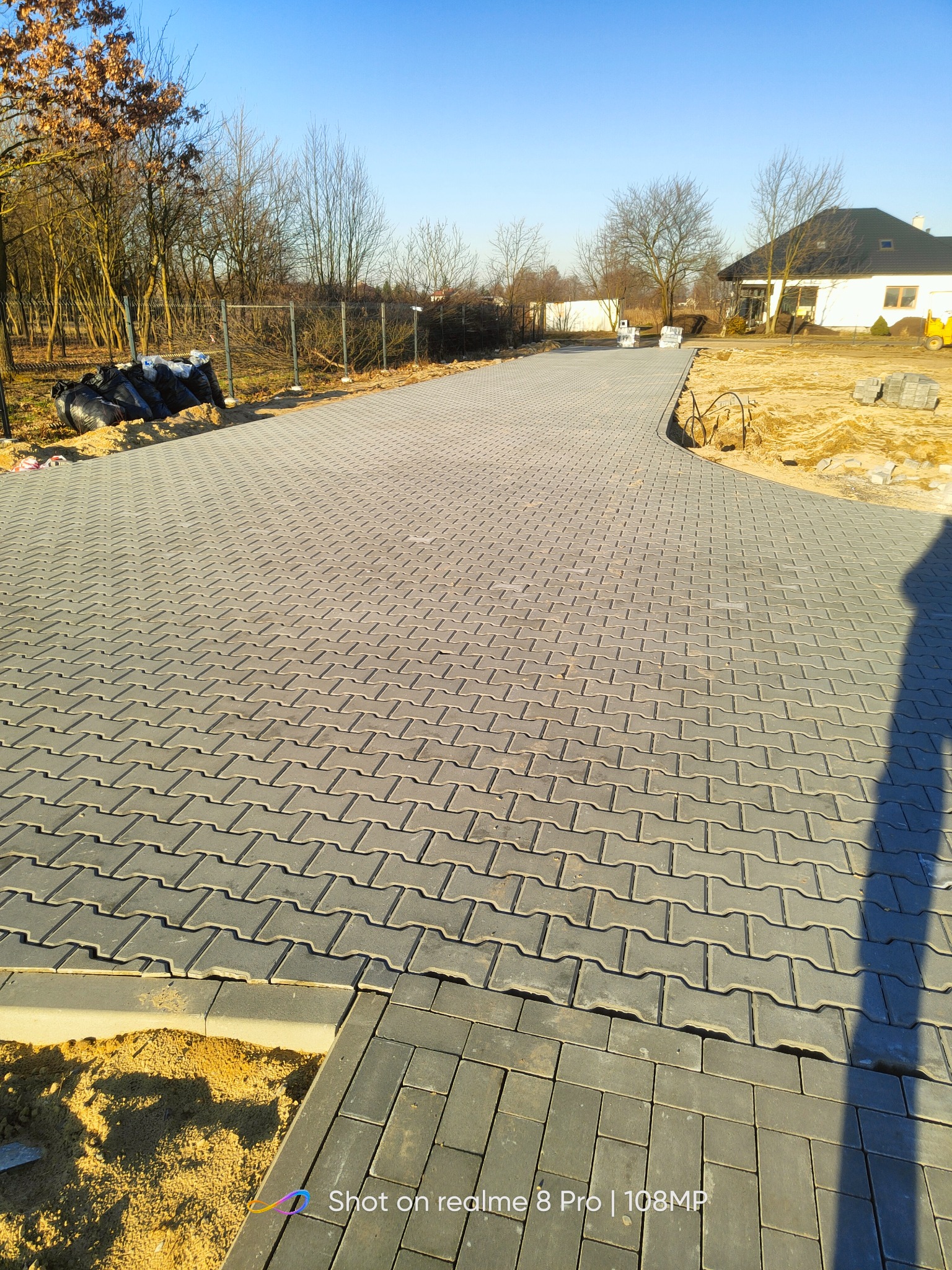 Ułożona szara kostka brukowa w kształcie litery H tworząca długi podjazd do domu jednorodzinnego w słoneczny dzień, widoczne obrzeża i przygotowane podłoże.