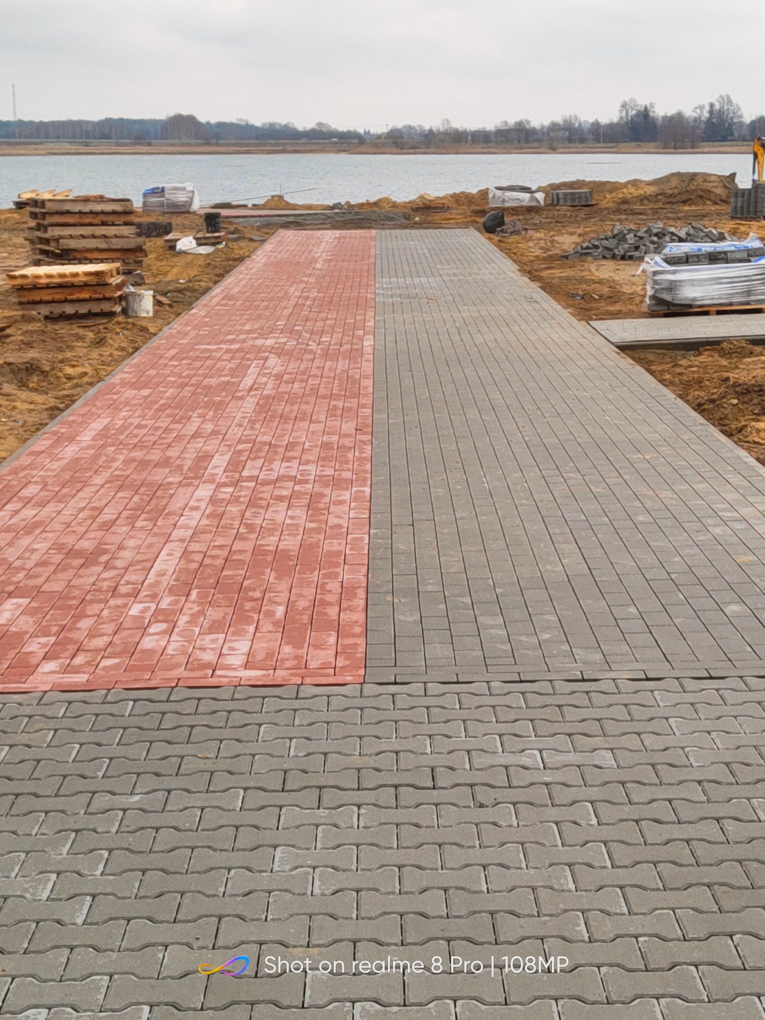 Ścieżka z kostki brukowej w dwóch kolorach, czerwonym i szarym, prowadząca w kierunku jeziora w pochmurny dzień. Palety i materiały budowlane widoczne po bokach.