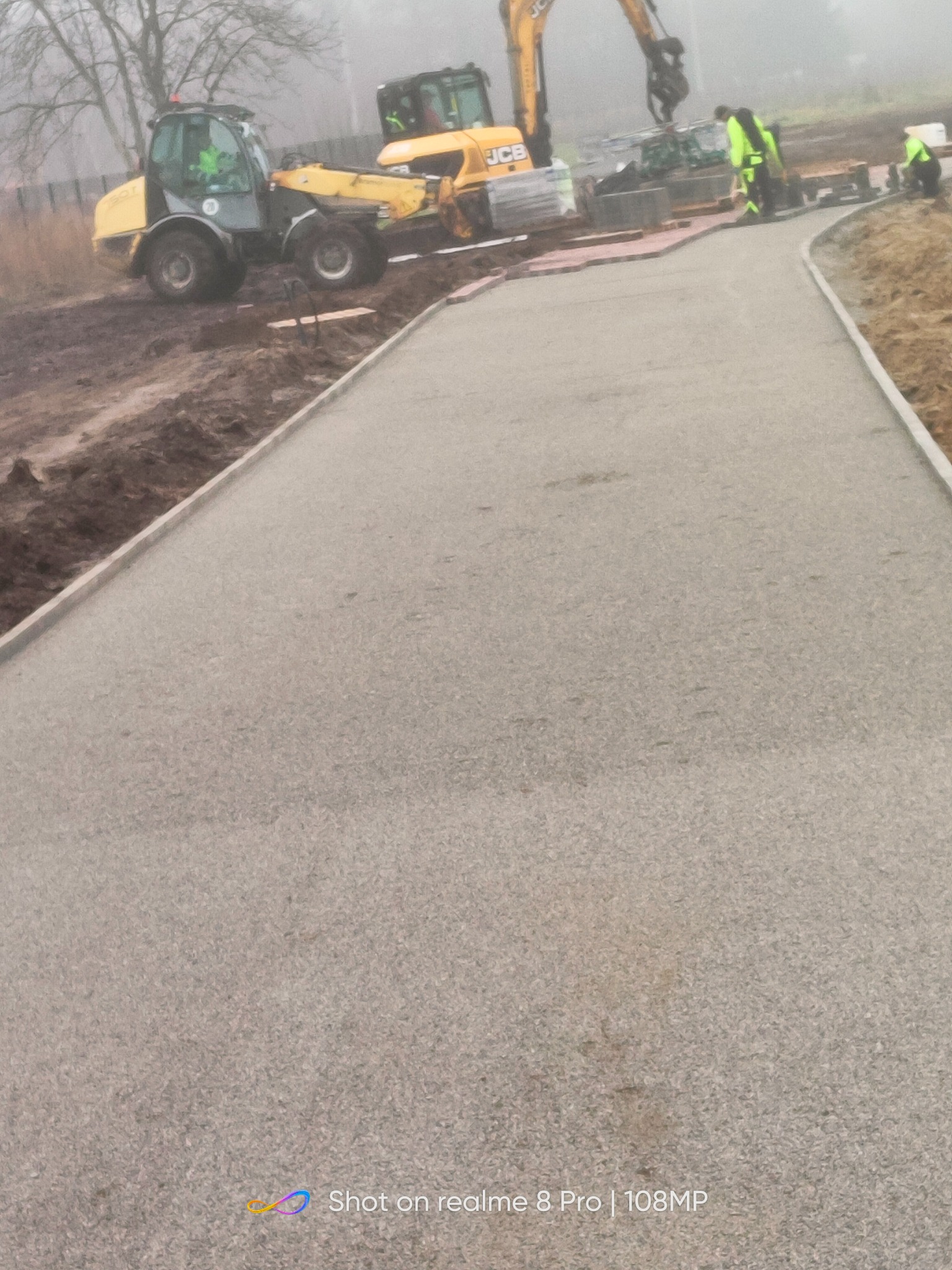 Budowa drogi: widok na nową, utwardzoną nawierzchnię z szarego kruszywa, obok pracujące maszyny budowlane JCB i robotnicy w odblaskowych kamizelkach. W tle drzewa we mgle.