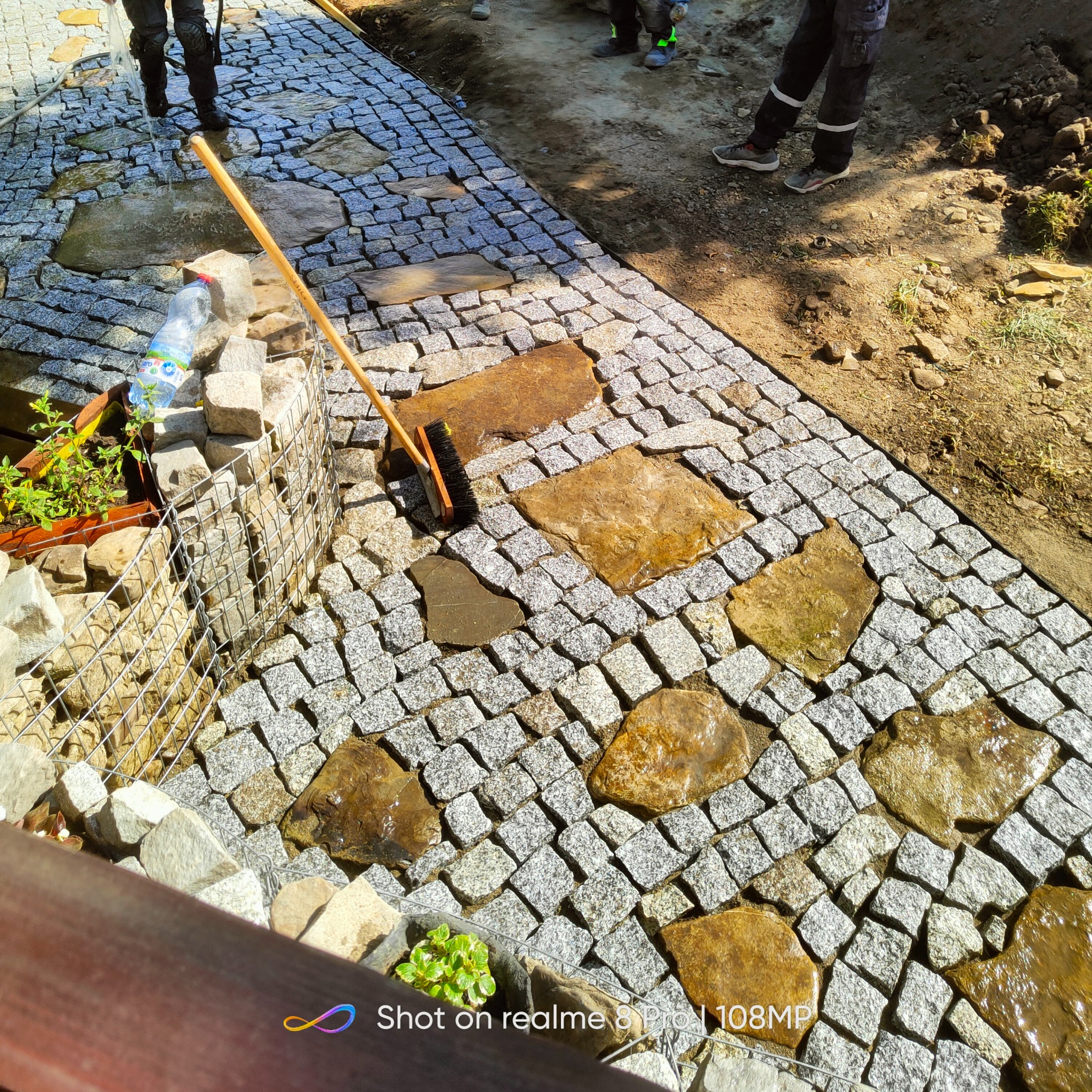 Świeżo ułożona ścieżka z kostki granitowej i nieregularnych płyt kamiennych, częściowo mokra, z widoczną szczotką do zamiatania, obok rabata z roślinami w drucianej siatce.