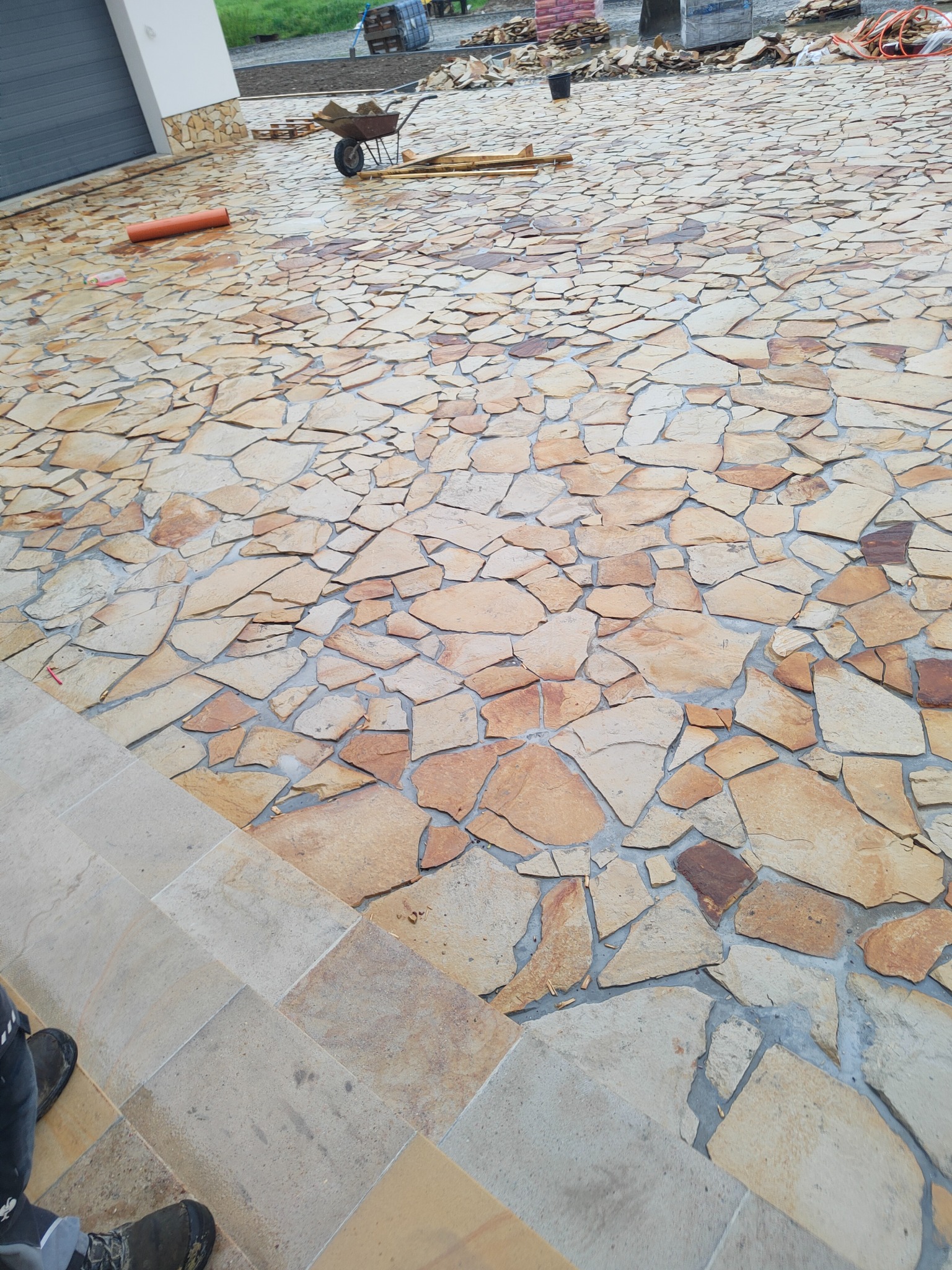 Nierówna nawierzchnia z nieregularnych, brązowo-beżowych kamieni, w tle taczka z materiałami budowlanymi i fragment budynku z szarą bramą garażową.