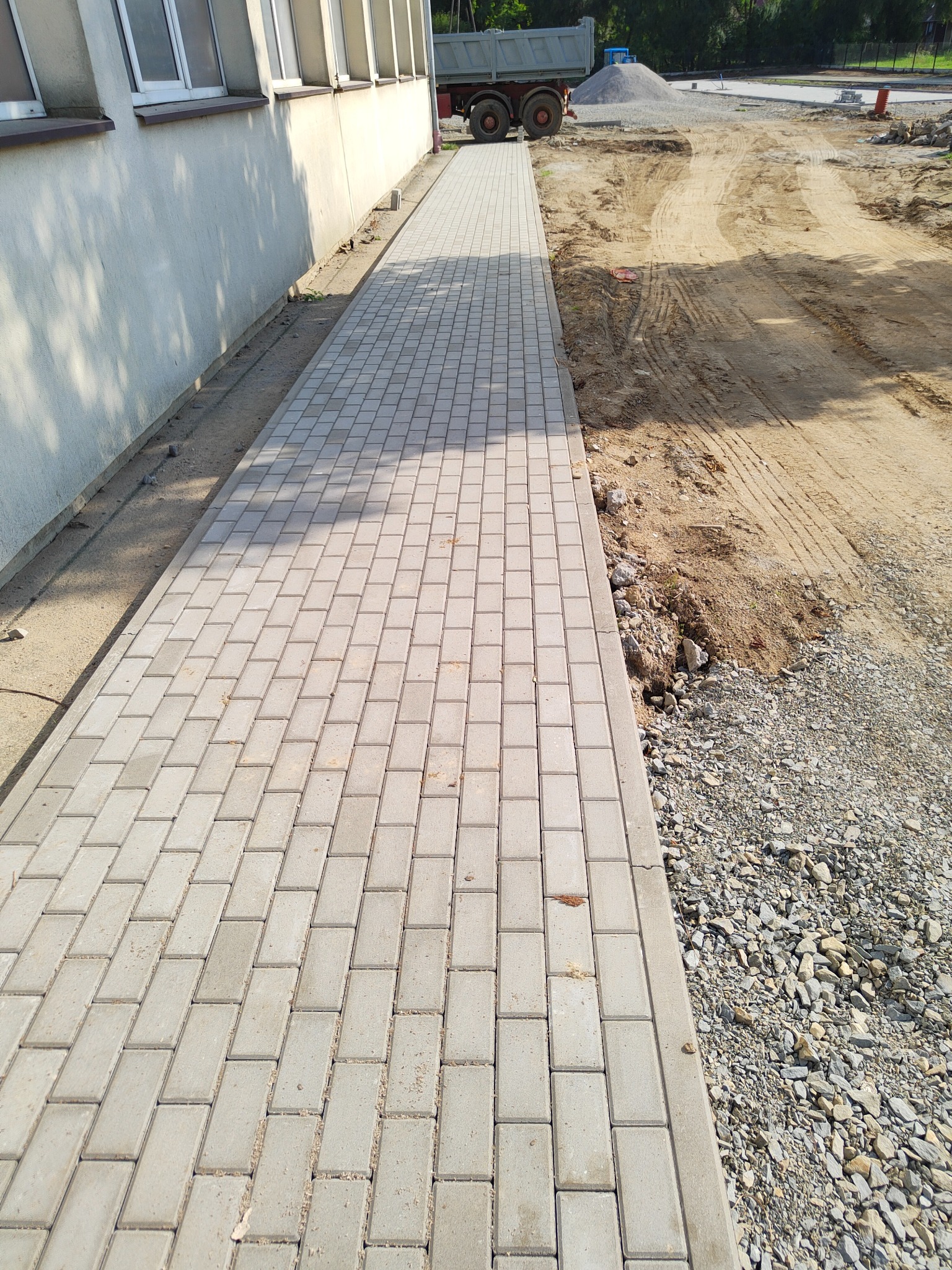 Ułożona, szara kostka brukowa tworząca chodnik wzdłuż budynku, z widocznymi śladami opon ciężarówki na ziemi obok.