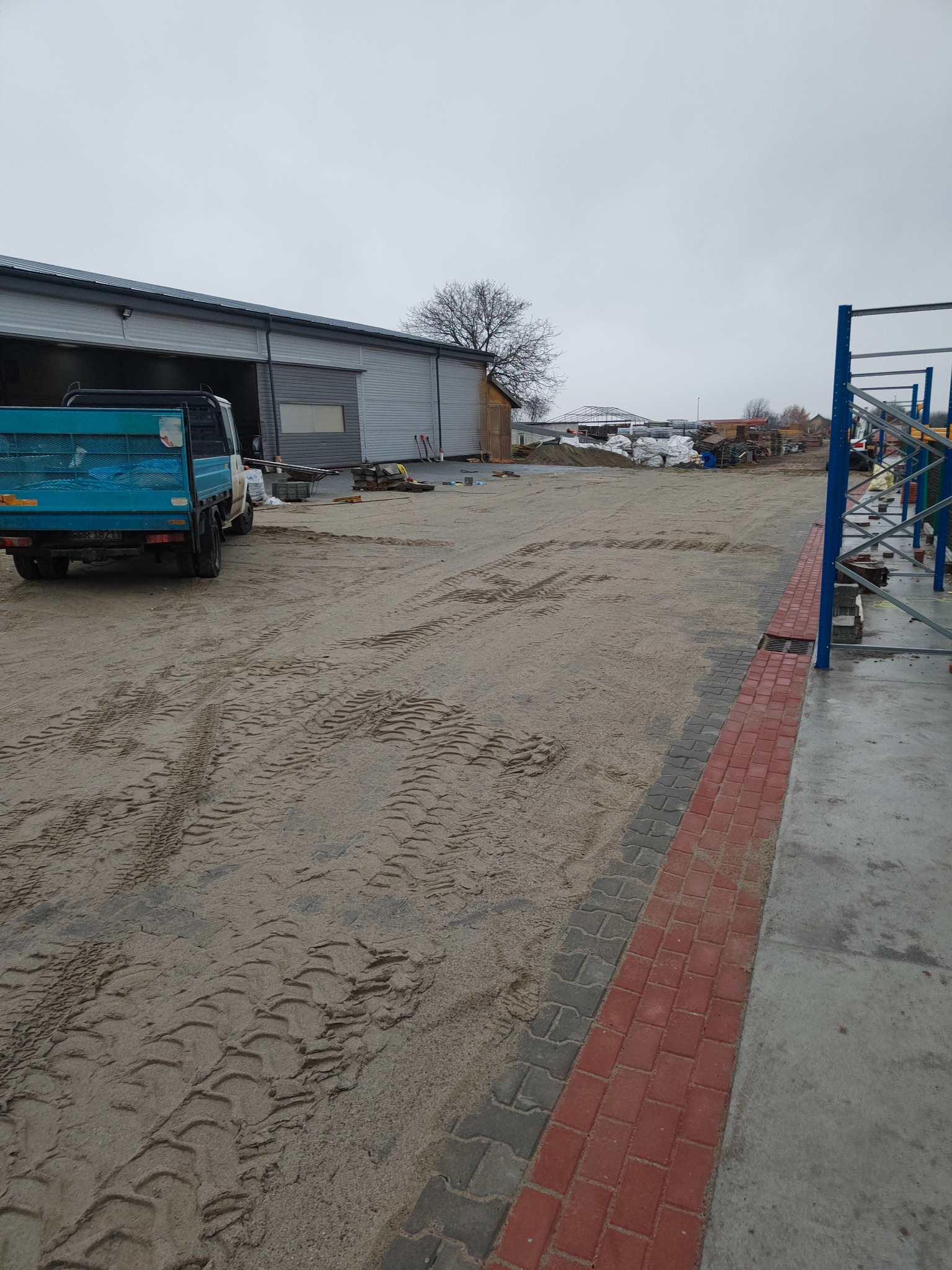 Teren budowy z widocznymi śladami opon ciężarówki na piasku, obok magazyn z otwartą bramą i częściowo ułożona nawierzchnia z czerwono-szarej kostki.
