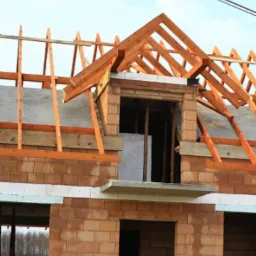 Surowy stan domu z czerwonej cegły ceramicznej z drewnianą konstrukcją dachu i lukarną w trakcie budowy, widoczne okno i otwór drzwiowy na parterze.