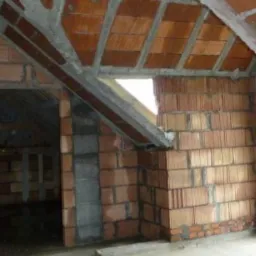 Wnętrze domu w trakcie budowy, widoczne ściany z czerwonej cegły i sufit z betonowymi wzmocnieniami, fragment okna dachowego, surowy stan wykończenia.