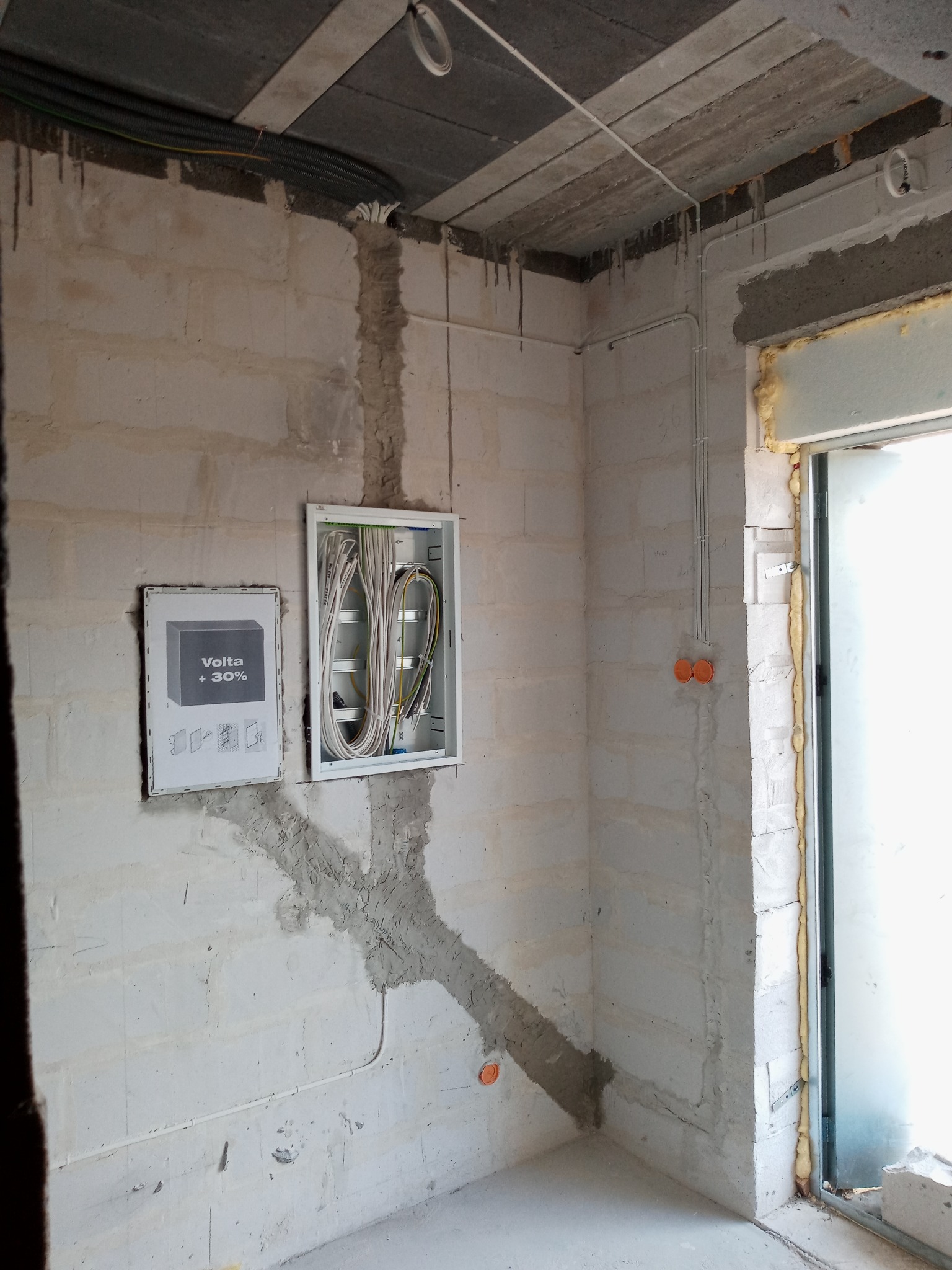 Surowy stan wnętrza w trakcie prac elektrycznych: otwarta skrzynka rozdzielcza z przewodami, bruzdy w ścianie z przewodami, puszki elektryczne, plakat informacyjny Volta +30% na ścianie z betonu...