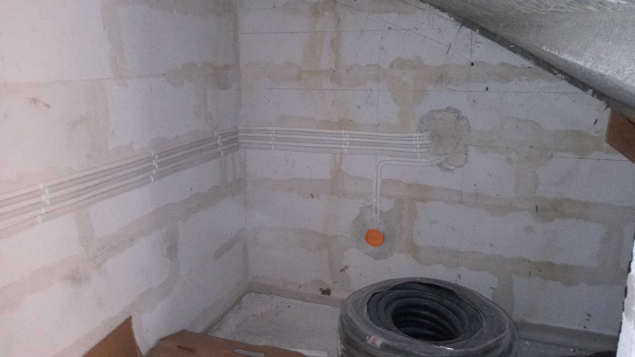 Białe kable elektryczne ułożone poziomo w rurkach na surowej ścianie z betonu komórkowego w trakcie montażu, widoczna rolka kabla w folii i pomarańczowa puszka instalacyjna.