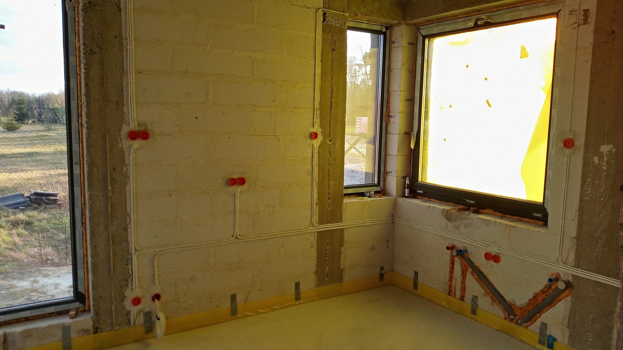 Surowy stan wnętrza z widocznymi przewodami elektrycznymi i instalacjami wodno-kanalizacyjnymi w ścianach z betonu komórkowego, okna zabezpieczone folią, widok na zewnątrz.