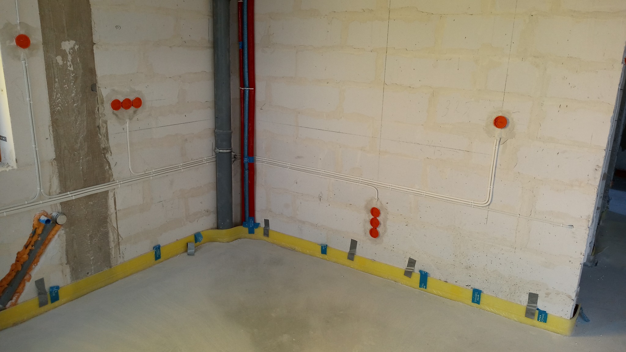 Surowy stan wykończeniowy pomieszczenia z widocznymi przewodami elektrycznymi w białych rurkach na ścianach z betonu komórkowego, pomarańczowymi puszkami instalacyjnymi, rurami kanalizacyjnymi...