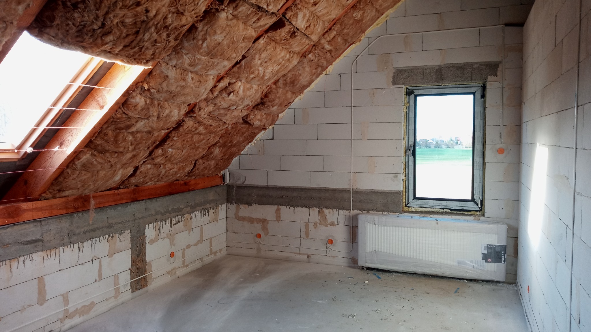 Poddasze w trakcie wykończenia: ocieplona wełną mineralną skośna ściana pod oknem dachowym, surowe ściany z betonu komórkowego z widocznymi przewodami elektrycznymi i nowym grzejnikiem w folii.