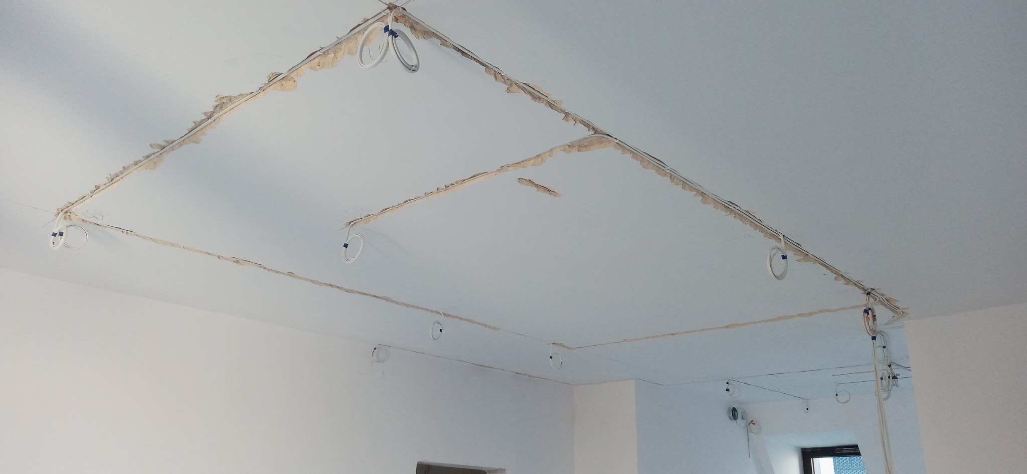 Układanie przewodów elektrycznych w bruzdach podtynkowych na suficie w trakcie remontu, widoczne przewody przytwierdzone do sufitu za pomocą niebieskiej taśmy.