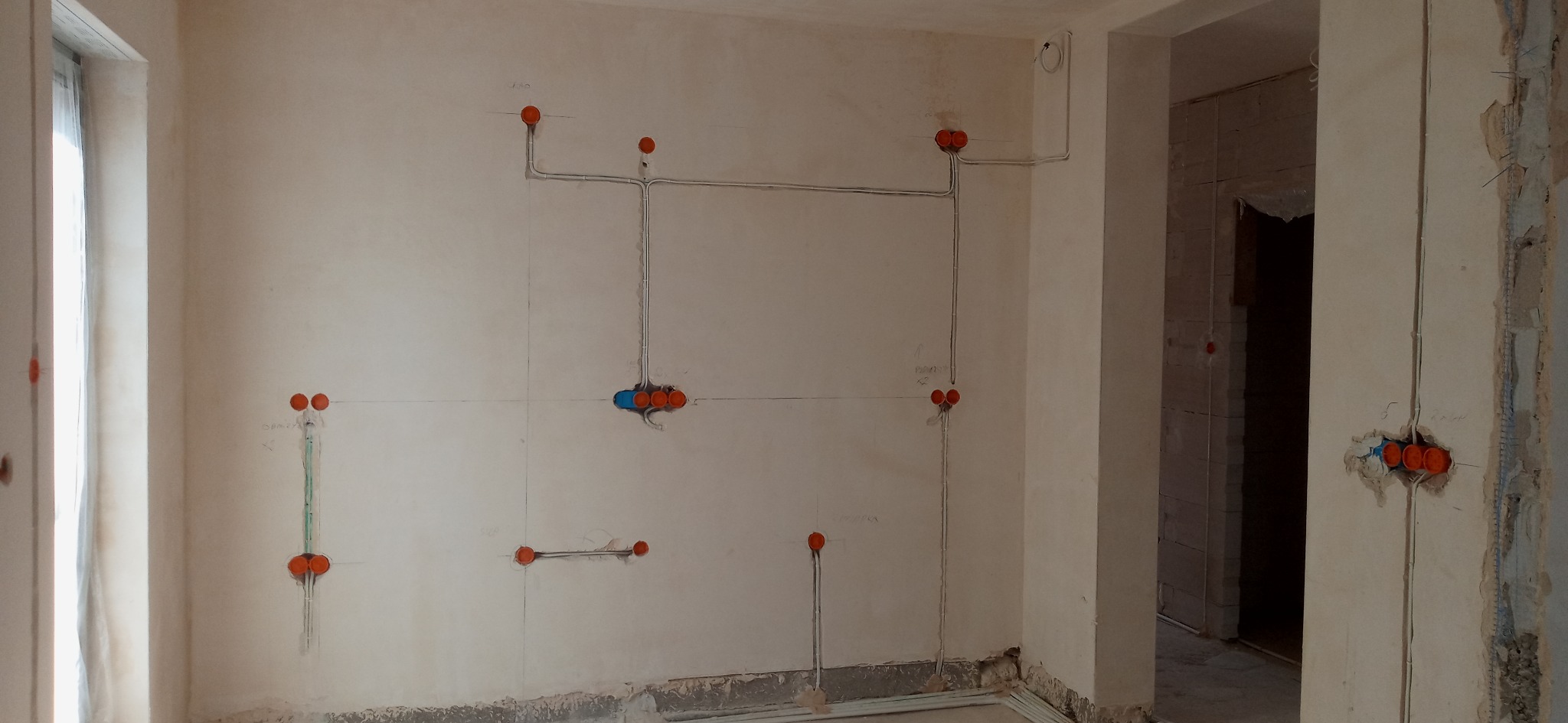 Wewnętrzne rozprowadzenie przewodów elektrycznych w surowym stanie, widoczne puszki instalacyjne z pomarańczowymi dekielkami i poprowadzone kable po ścianach z naniesionymi oznaczeniami ołówkiem.