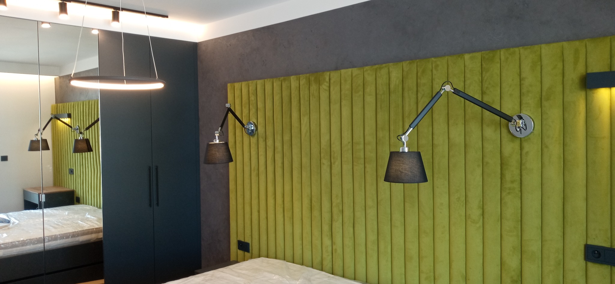 Sypialnia z zielonym, żebrowanym panelem ściennym, regulowanymi lampami ściennymi z czarnymi abażurami, ciemnoszarą ścianą i szafą w zabudowie, widoczne gniazdko elektryczne.