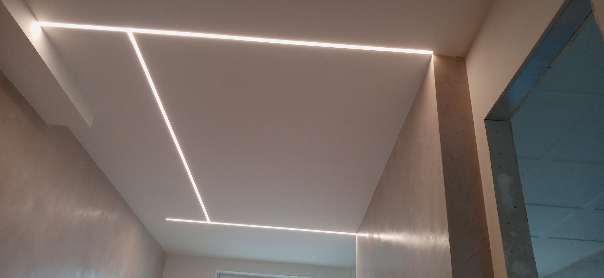 Nowoczesne oświetlenie sufitowe z liniowymi lampami LED tworzącymi geometryczny wzór w minimalistycznym wnętrzu z dekoracyjnym tynkiem na ścianach.