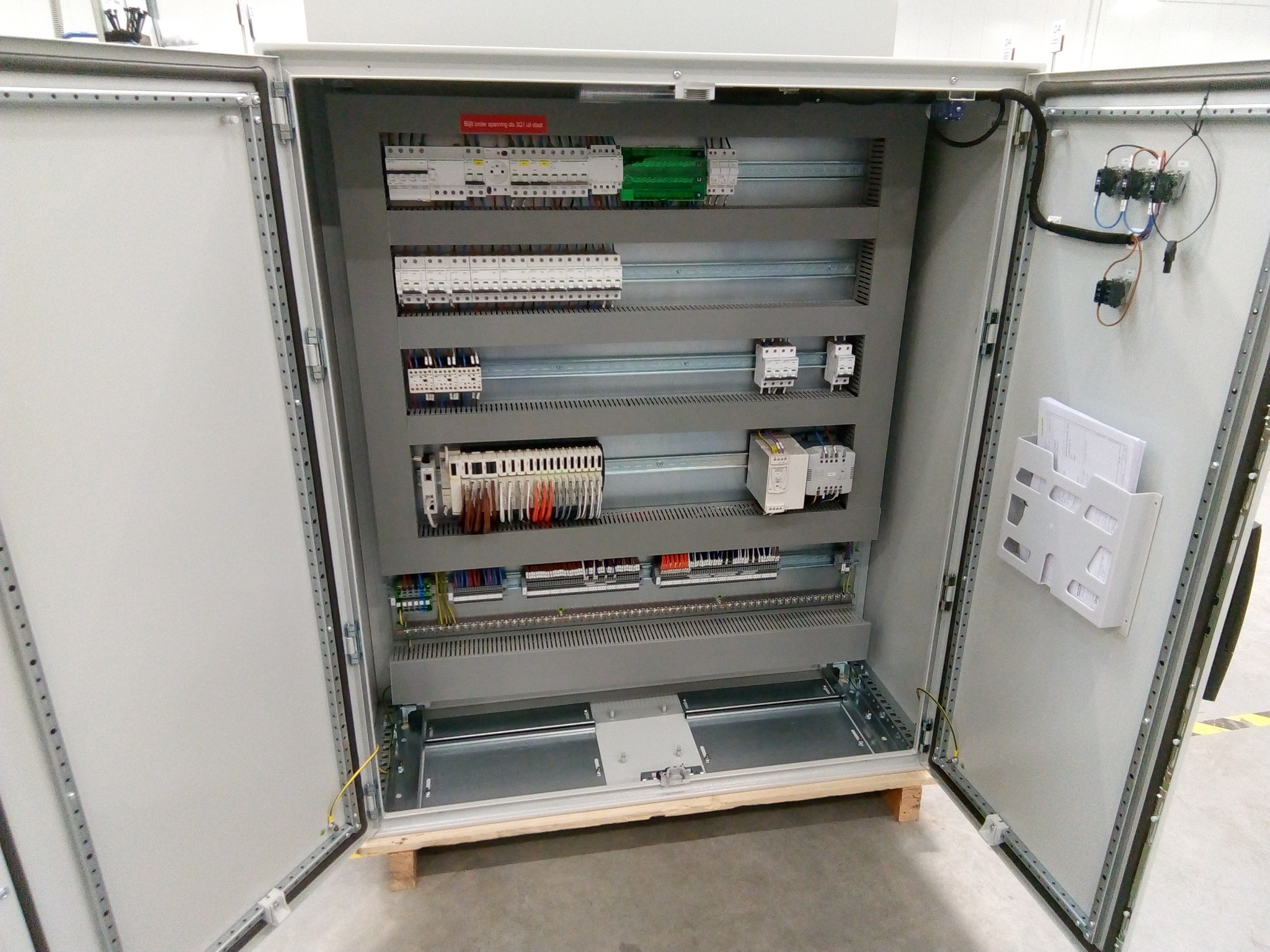 Otwarta szafa sterownicza z widocznymi elementami instalacji elektrycznej, w tym bezpieczniki, listwy zaciskowe i przewody, zamontowana na drewnianej palecie.