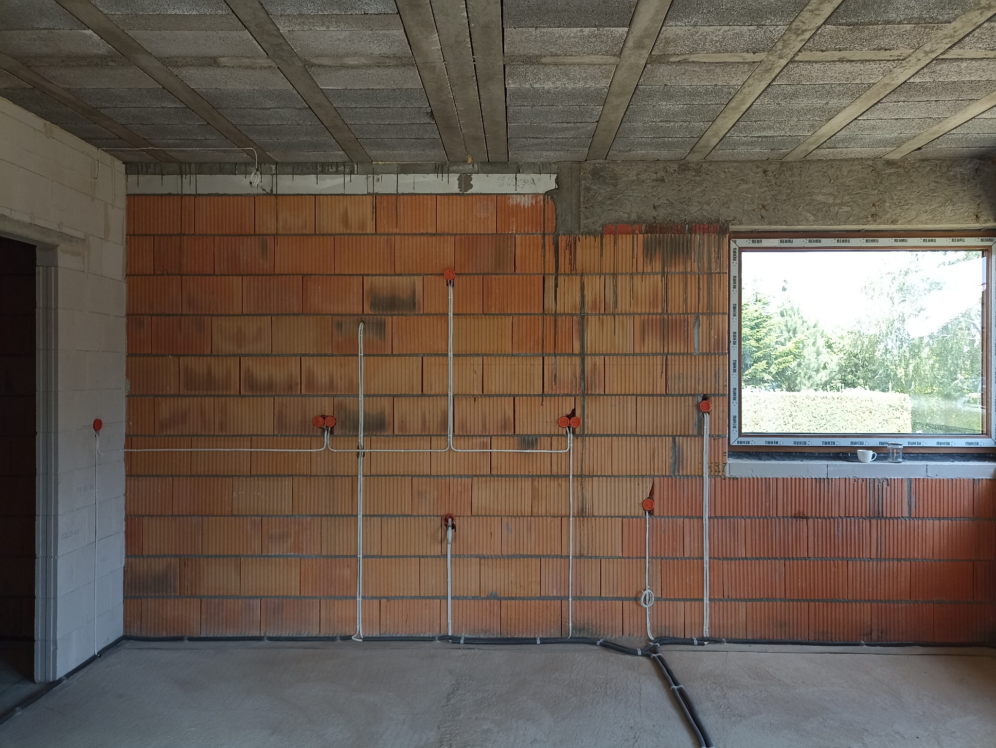 Surowy stan wnętrza z instalacją elektryczną: pomarańczowe puszki podtynkowe i białe rurki prowadzące kable na ceglanej ścianie, widok na okno z parapetem, betonowa wylewka i sufit.