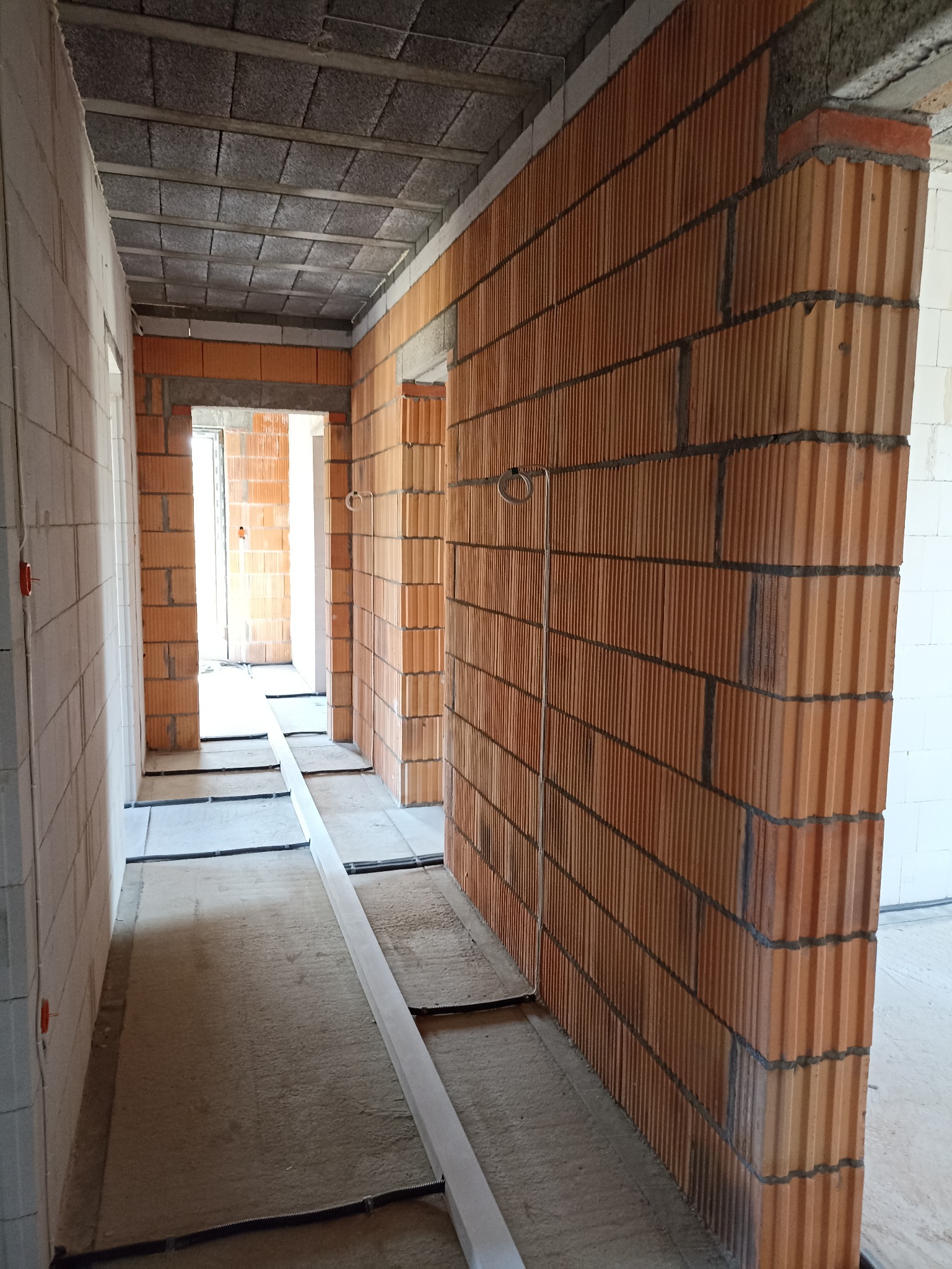 Wnętrze w trakcie budowy: widok na korytarz z ceglanymi ścianami, sufitem z betonowych płyt i elementami instalacji elektrycznej w podłodze.