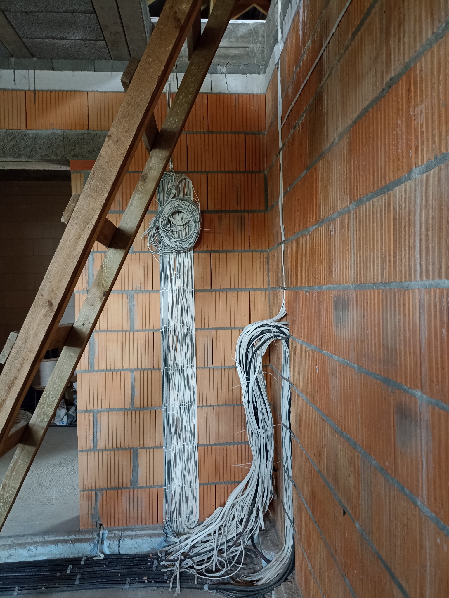 Surowe ściany z cegły dziurawki w trakcie prac elektrycznych, kable prowadzone w pionowych bruzdach, zwój kabli przymocowany do ściany, drewniana drabina oparta o ścianę, widok na surowy strop.