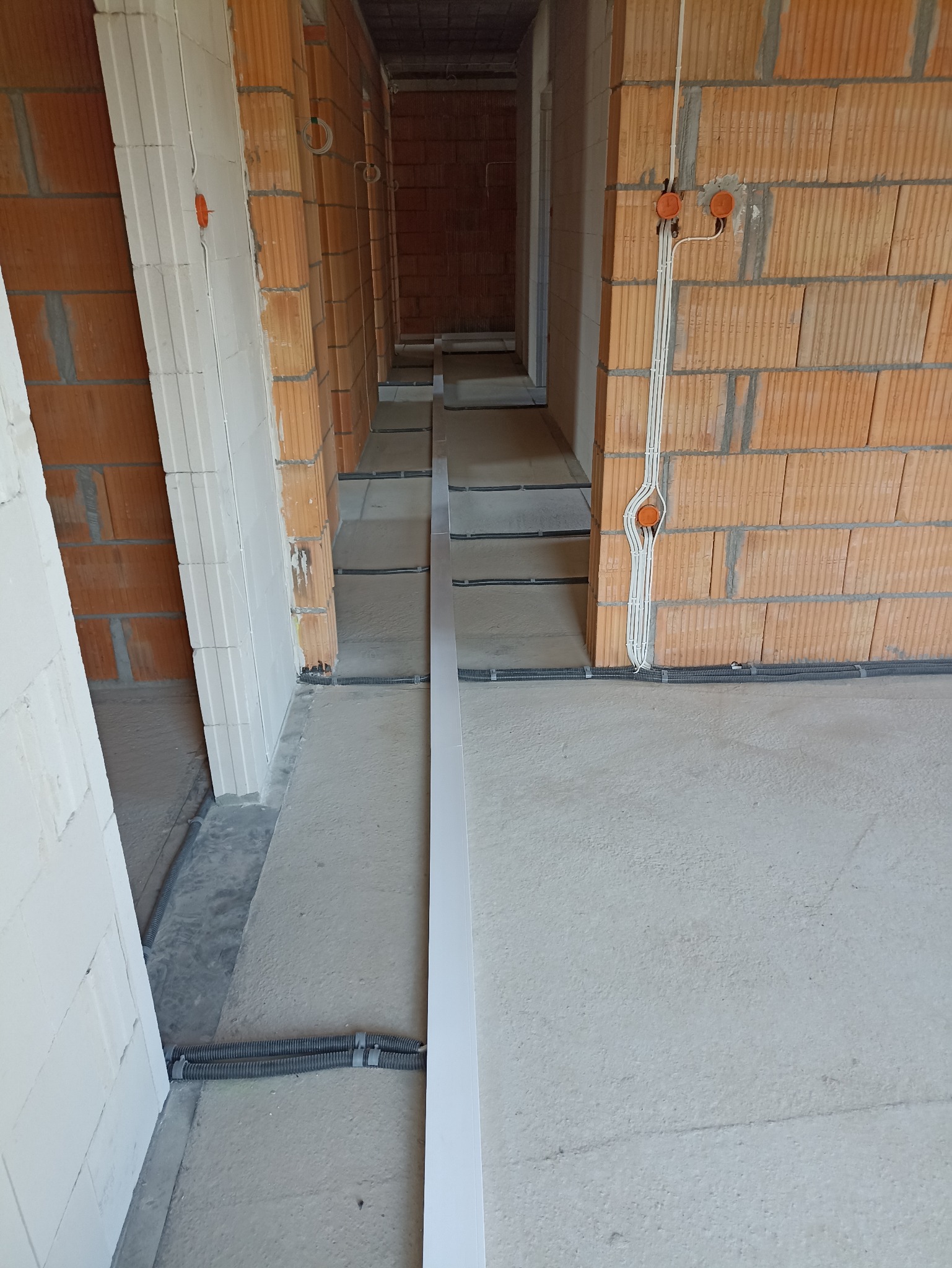 Wnętrze w stanie surowym z widocznymi przewodami elektrycznymi w peszlach na podłodze i wyprowadzonymi kablami w ścianach z czerwonej cegły.