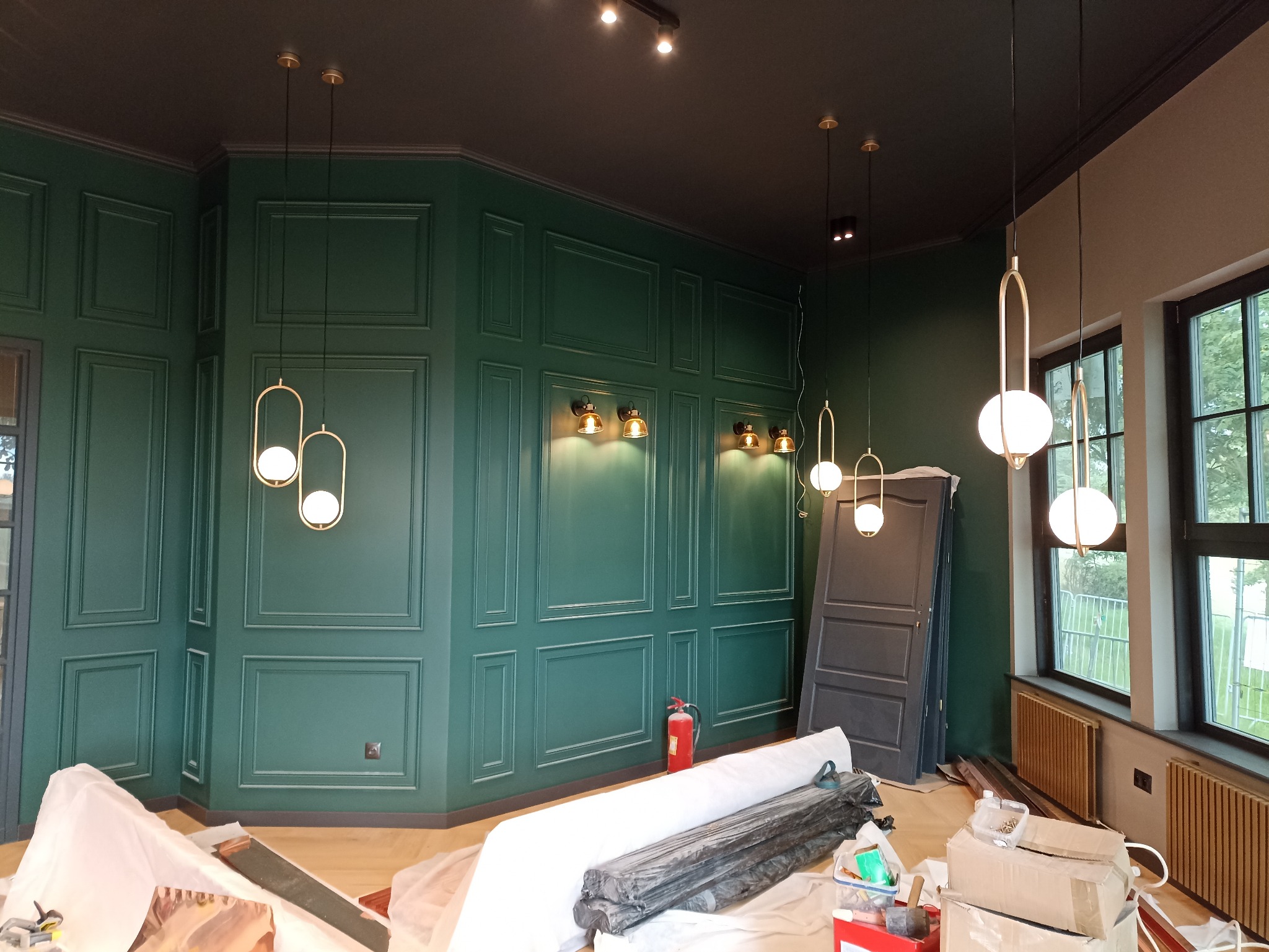Wnętrze w trakcie wykańczania: zielone ściany z dekoracyjnymi panelami, wiszące lampy kuliste i ścienne kinkiety, widoczne elementy instalacji elektrycznej i przygotowania do montażu, podłoga...