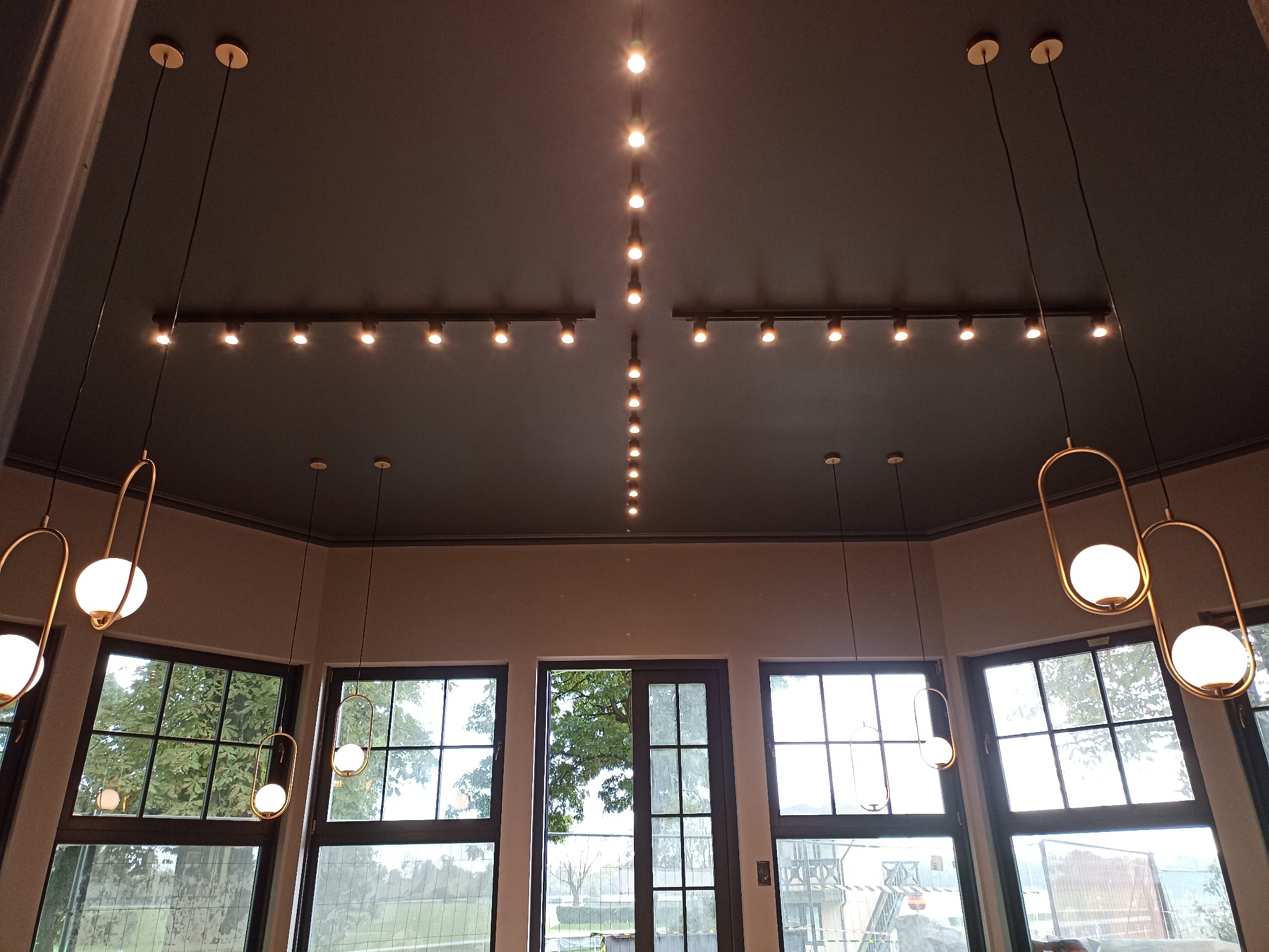 Nowoczesne oświetlenie sufitowe w pomieszczeniu z dużymi oknami: kombinacja szynoprzewodów z reflektorami i wiszących lamp kulistych z metalowymi oprawami.