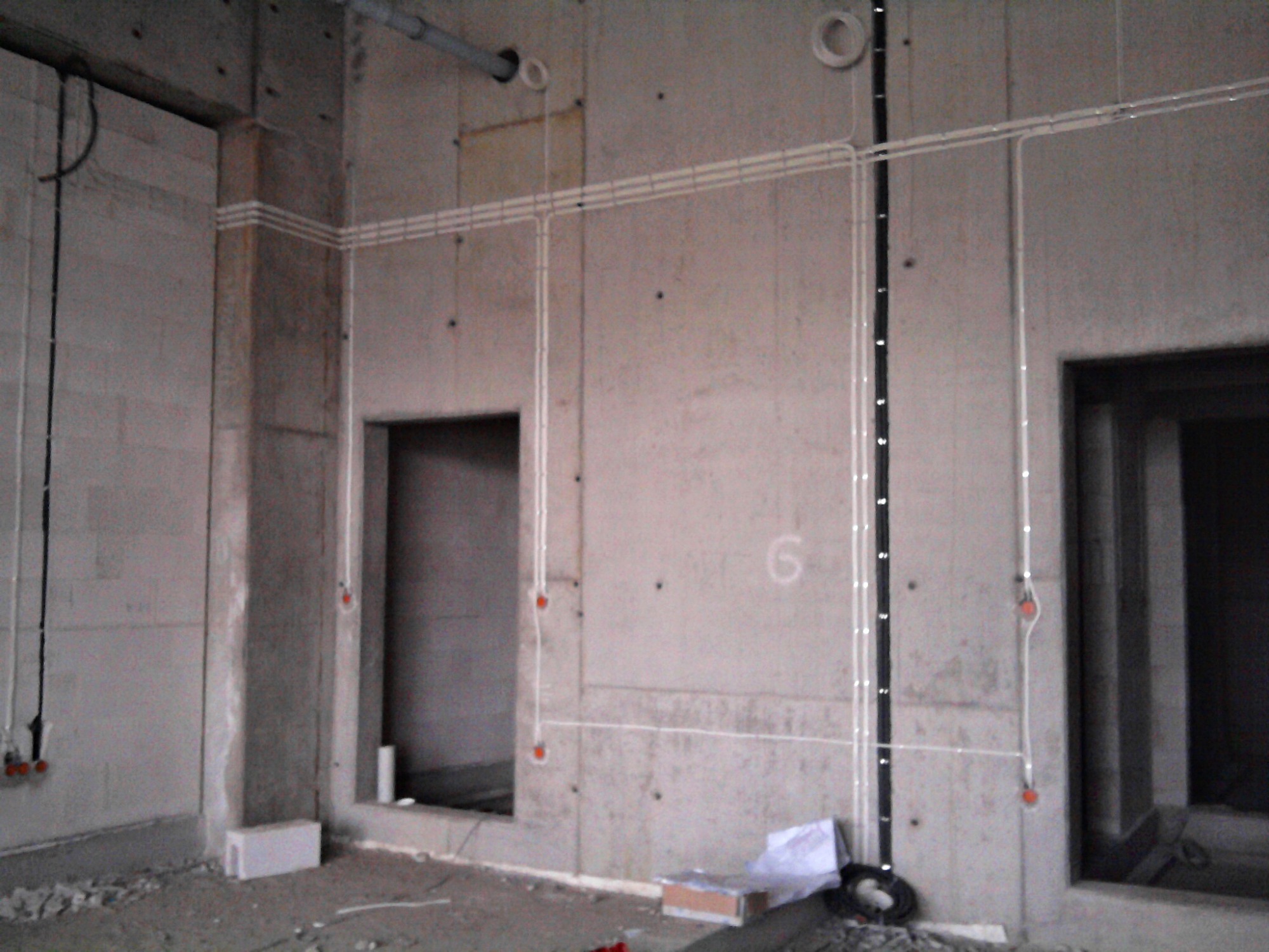 Surowa ściana betonowa z widocznymi przewodami elektrycznymi ułożonymi w peszlach oraz kablami w czarnej listwie, w tle otwory drzwiowe, na podłodze gruz i zwinięty kabel.