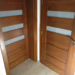 Dwa skrzydła drzwiowe z drewna o ciepłym odcieniu brązu, z poziomymi przeszkleniami, widoczne okucia i zawiasy, na podłodze panele imitujące drewno.