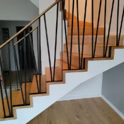 Nowoczesne schody z drewnianymi stopniami, białymi podstopnicami i czarną, geometryczną balustradą, widok z dołu.