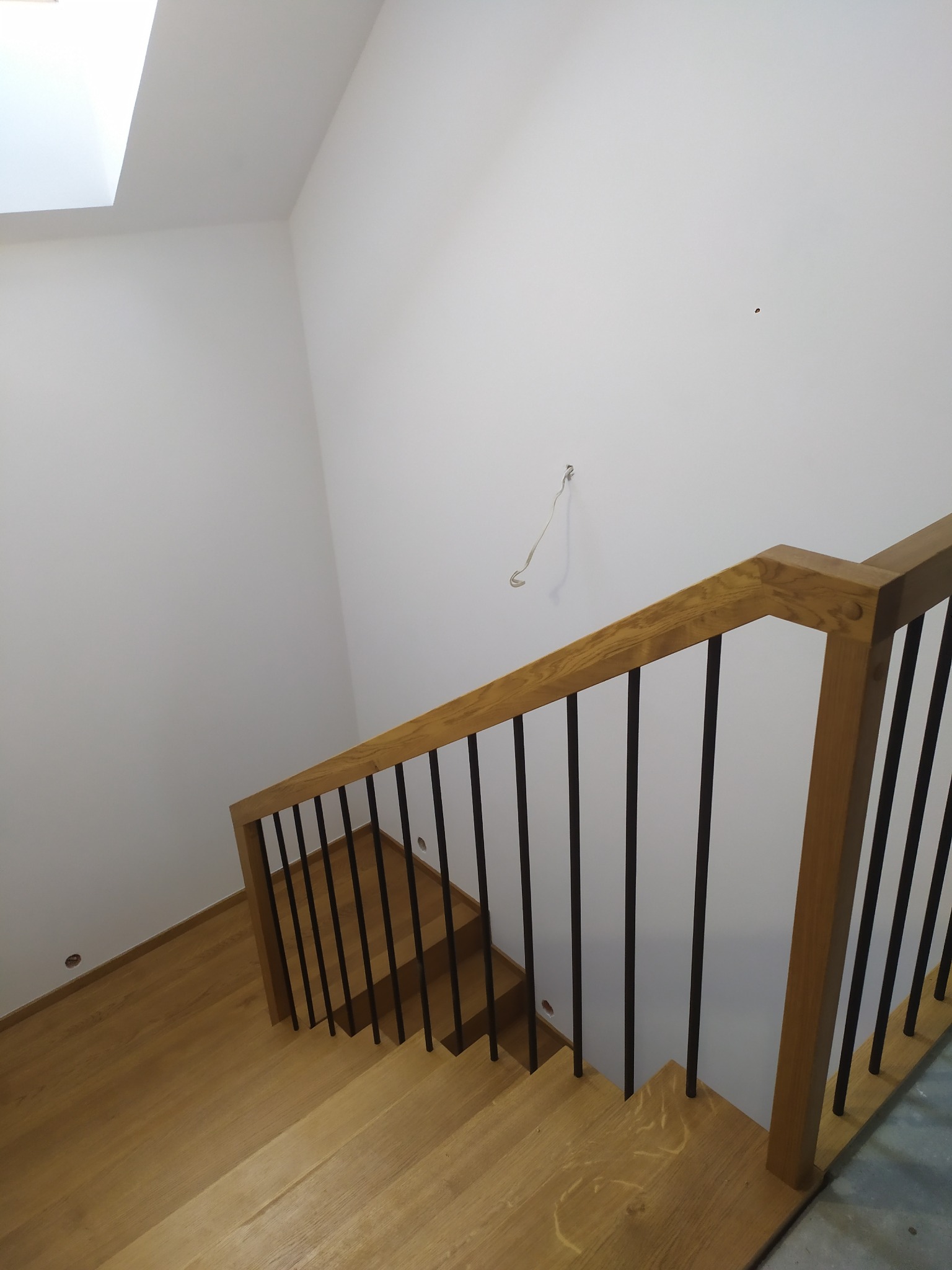 Widok z góry na jasne, dębowe schody z czarnymi, metalowymi balustradami, prowadzące w dół w nowoczesnym wnętrzu z białymi ścianami i świetlikiem.