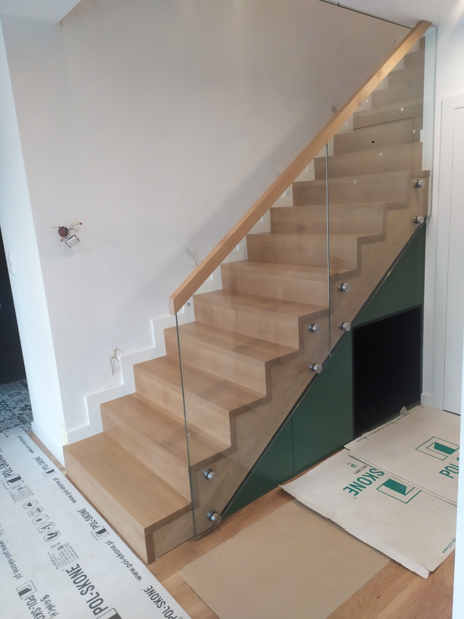 Nowoczesne schody drewniane z jasnego drewna, z balustradą ze szkła mocowaną na metalowych uchwytach, z drewnianą poręczą. Pod schodami zabudowa w kolorze ciemnej zieleni. Na podłodze...