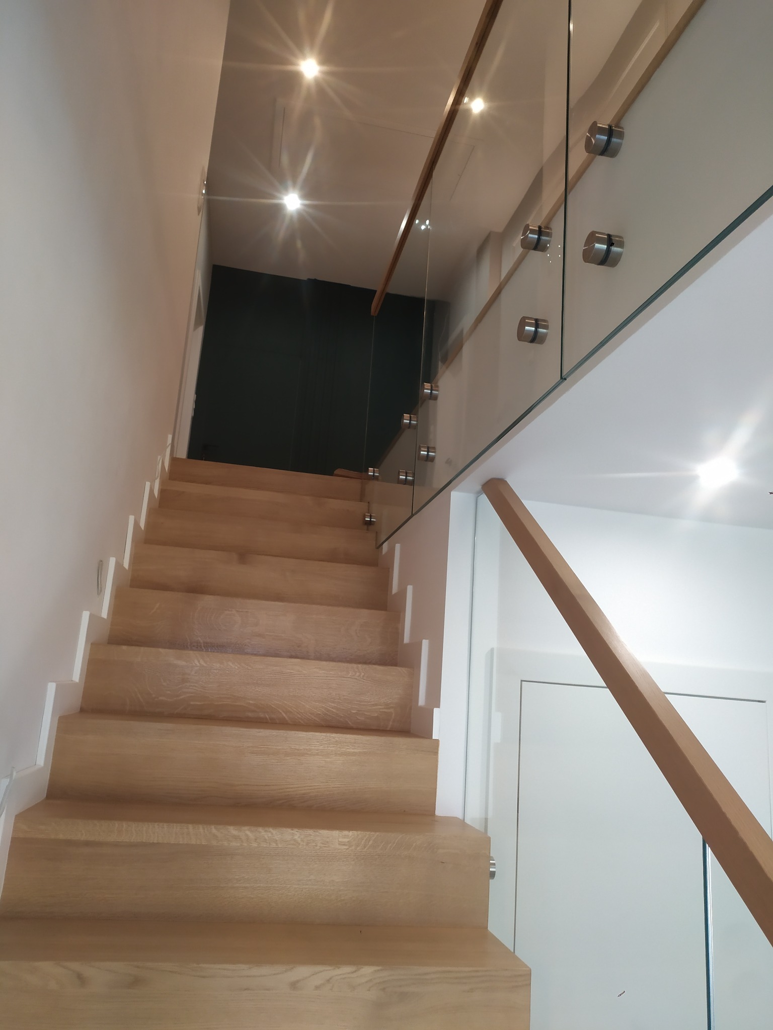 Jasne, dębowe stopnie schodów z białymi podstopnicami, zabezpieczone szklaną balustradą z drewnianą poręczą, mocowaną za pomocą metalowych uchwytów punktowych.