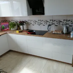 Biała, nowoczesna kuchnia z drewnianym blatem, na którym znajdują się różne akcesoria kuchenne, w tym patelnia na płycie indukcyjnej, akwarium i kwiaty.