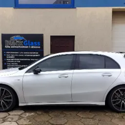 Biały Mercedes A-klasa z przyciemnionymi szybami bocznymi, zaparkowany przed budynkiem z reklamą firmy oferującej folie samochodowe i okienne.