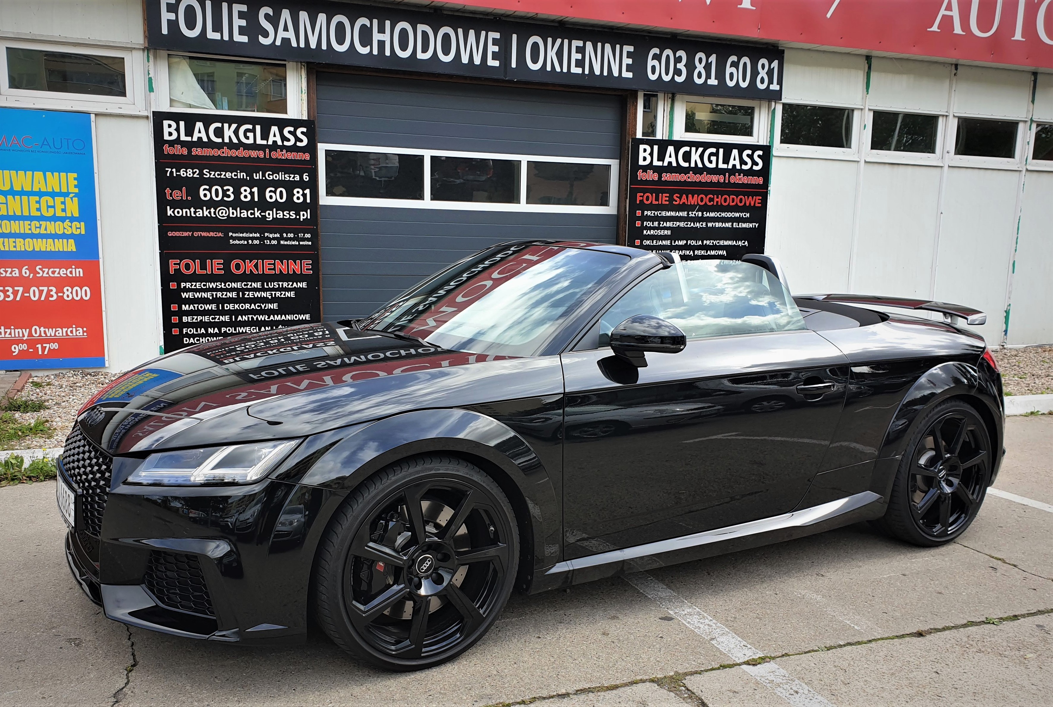 Czarny kabriolet Audi TT zaparkowany przed warsztatem Blackglass, z widocznymi reklamami folii samochodowych i okiennych, w tle szara brama garażowa.