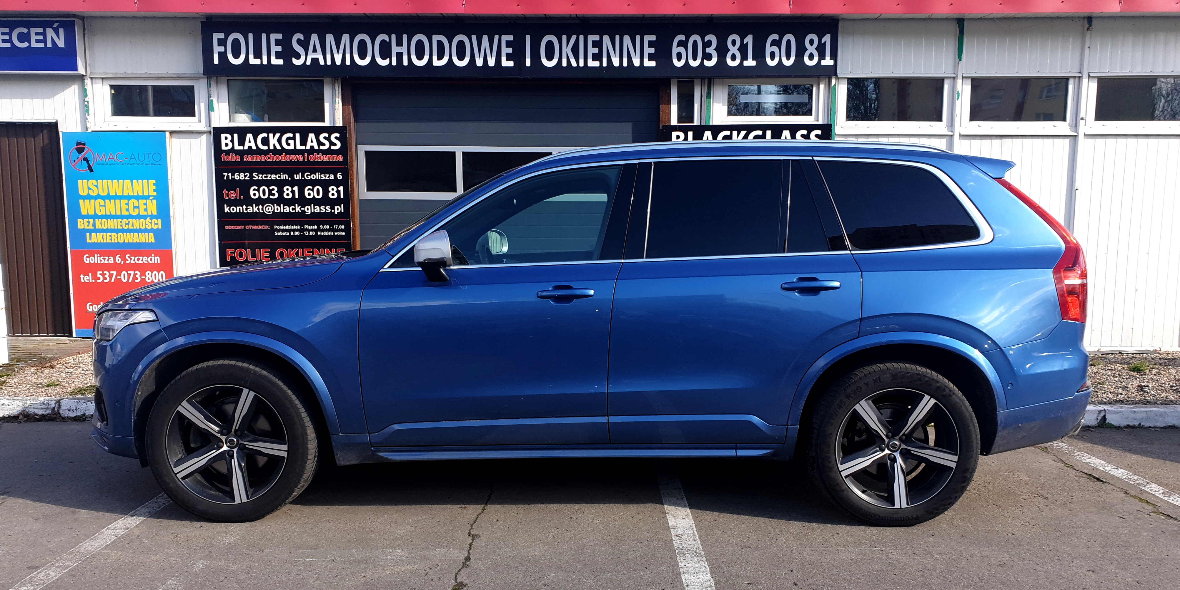 Niebieski SUV marki Volvo z przyciemnionymi szybami bocznymi, zaparkowany przed budynkiem firmy Blackglass oferującej folie samochodowe i okienne w Szczecinie, widoczny numer telefonu i adres e-mail.