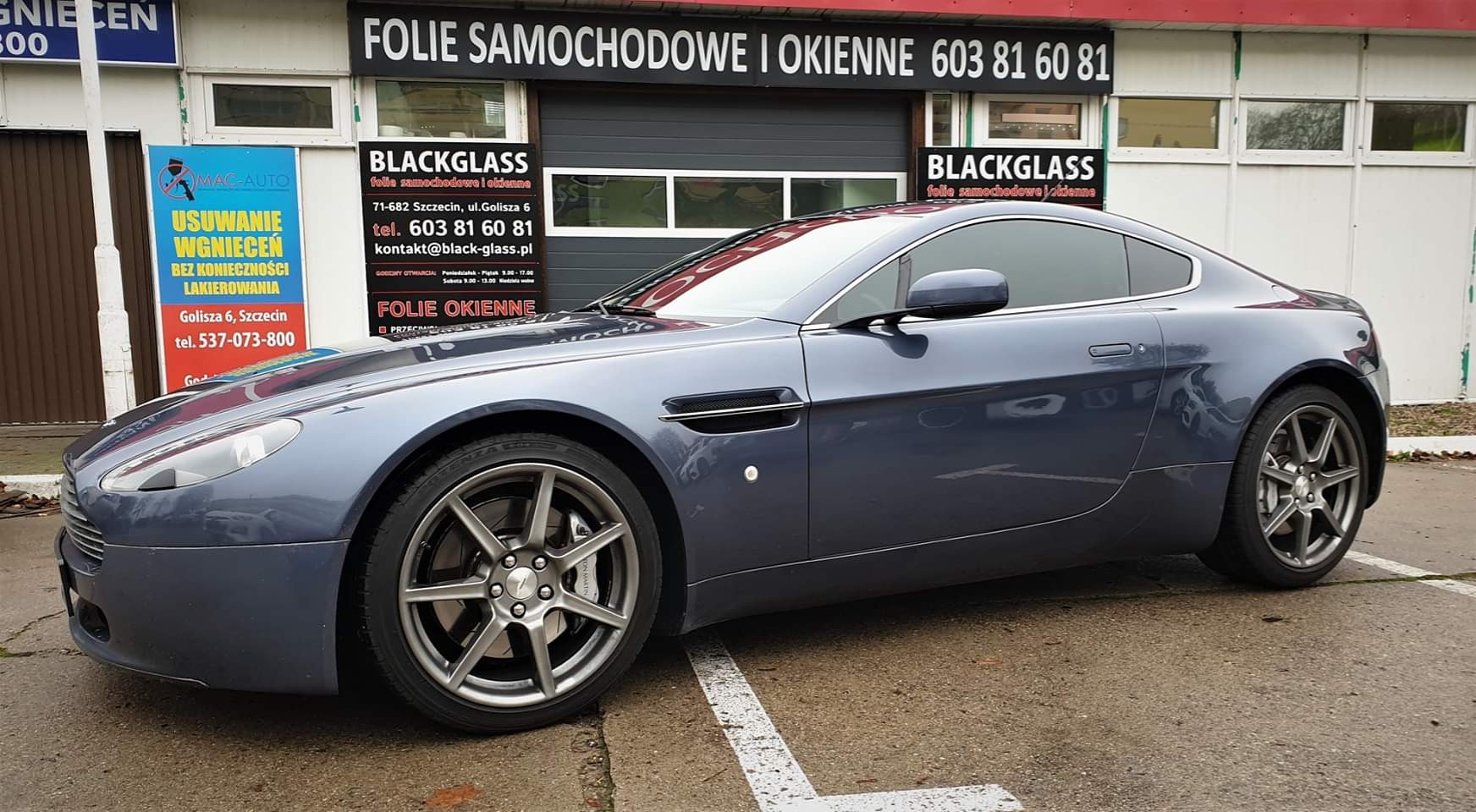 Granatowy Aston Martin z przyciemnionymi szybami zaparkowany przed warsztatem Blackglass w Szczecinie, z widocznymi reklamami usług folii samochodowych i okiennych.