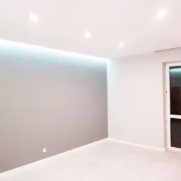 Wnętrze pokoju po remoncie: szara ściana z podświetleniem LED przy suficie, biała podłoga i białe drzwi balkonowe.